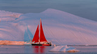 Red sail boat sailing past Iceberg in the midnight sun.