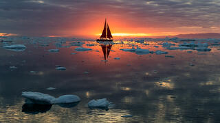 Red sail boat passing sun on horizon on Greenland waters.