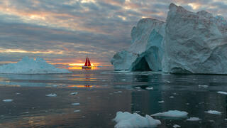 Red sail boat passing iceberg in Greenland waters.