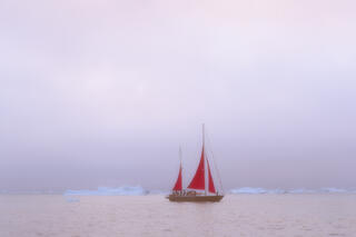 Red sail boat passing through the fog on Greenland waters.