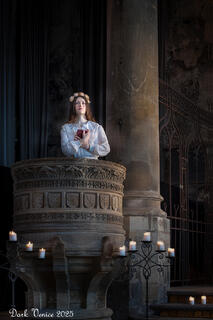 Female actor in costume in pulpit with book in hand looking up. 