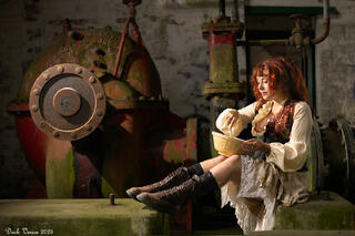 Female model in costume, sitting in disused pumping station stirring a mixture in a bowl.